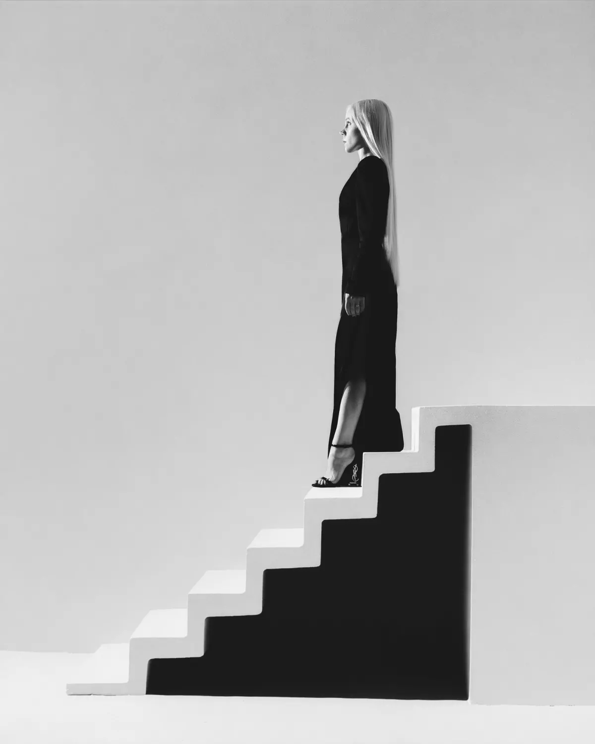 Чёрно-белый кадр: женщина в длинном тёмном платье, с очень длинными светлыми волосами, стоит в профиль на белой лестнице.
