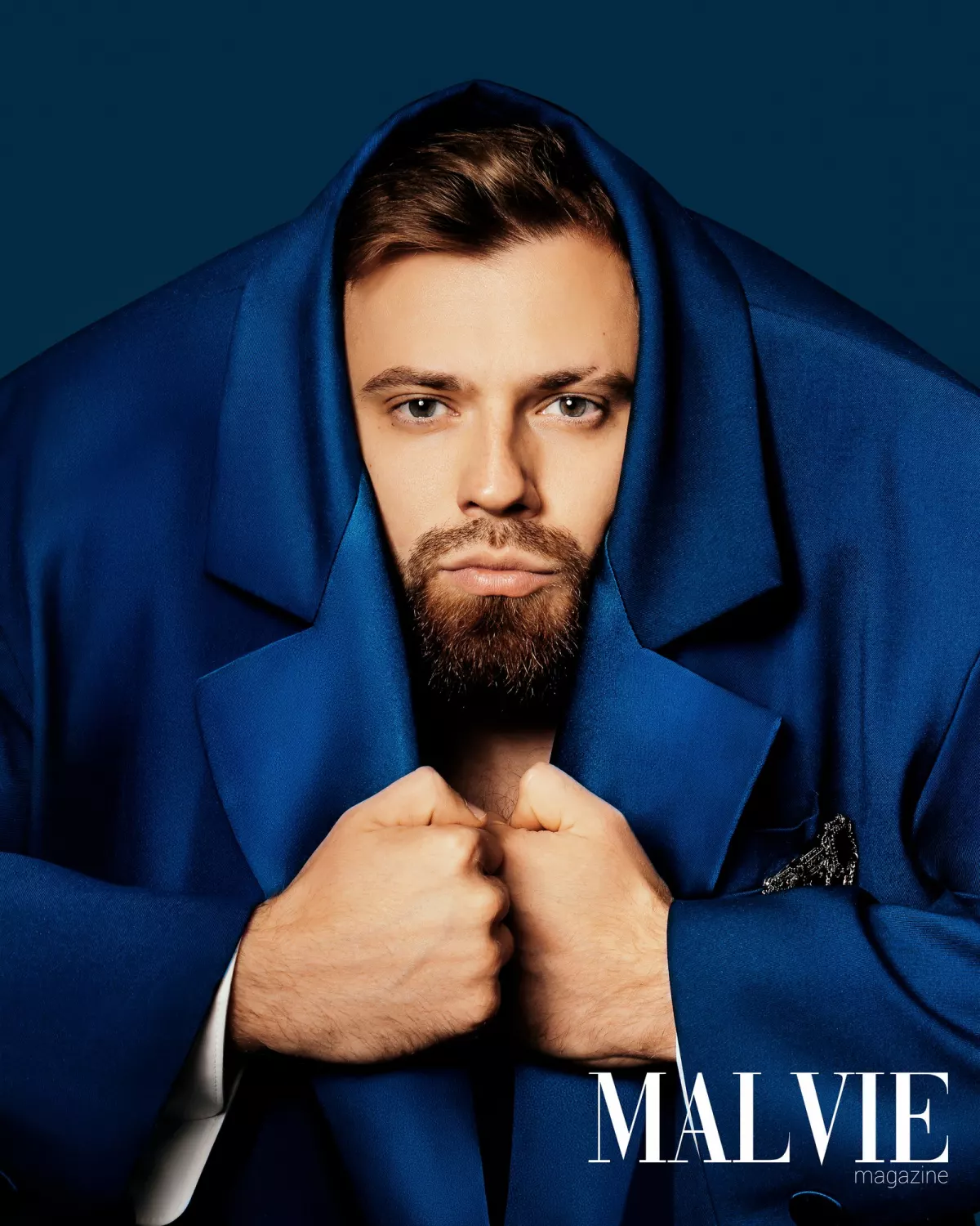 Портрет в журнале MALVIE: бородатый мужчина в ярко-синем пиджаке, накинутом как капюшон, сжимает лацканы кулаками; внизу справа логотип издания.